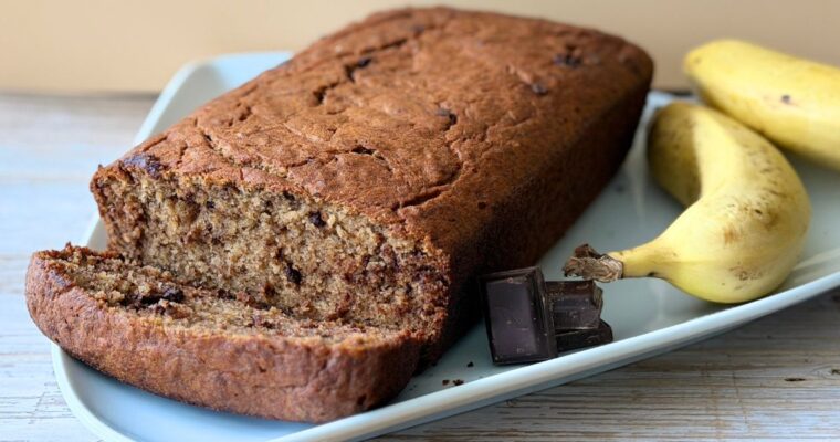 A Truly Yummy Banana Bread served on a white serving plate alongside yellow bananas and dark chocolate for display.