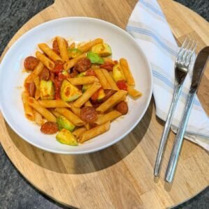 A single serving of chorizo & avocado pasta in a spicy tomato sauce on a serving board with napkin and cutlery